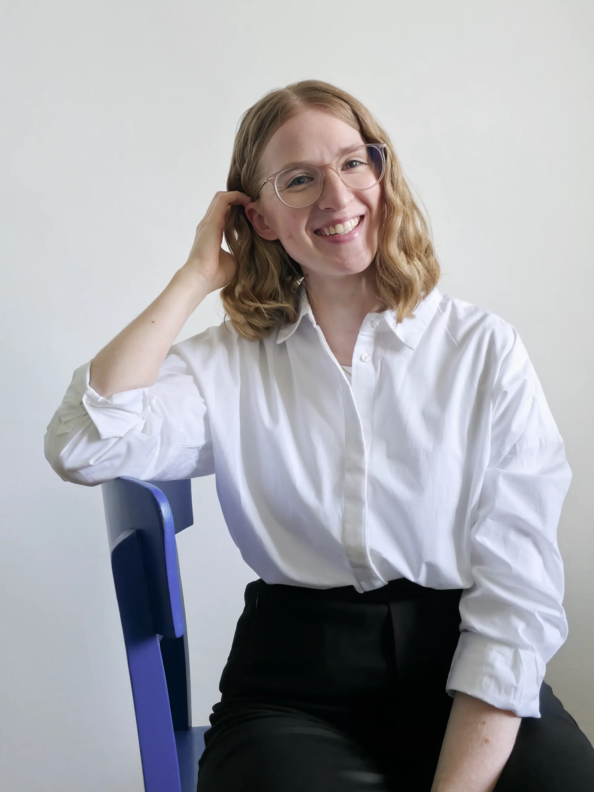 Anika Schulze, Freelance Grafikdesignerin aus Hamburg. Sie hat schulterlanges, lockiges, blondes Haar und trägt eine rosa, durchsichtige Brille. Sie steht vor einer wie-en Wand und trägt eine wie-e Hemdbluse und eine schwarze Anzughose.
