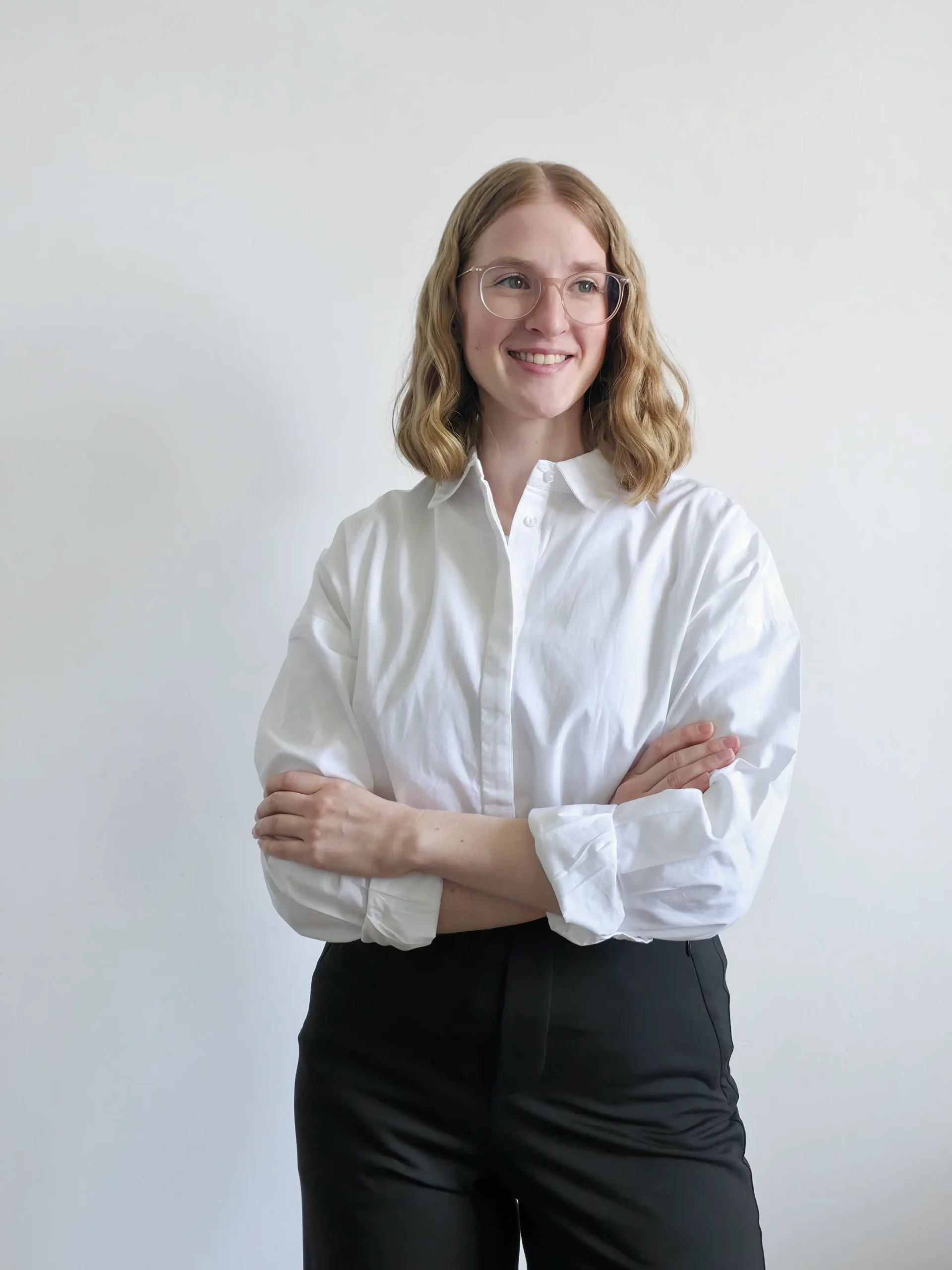 Anika Schulze, Freelance Grafikdesignerin aus Hamburg. Sie hat schulterlanges, lockiges, blondes Haar und trägt eine rosa, durchsichtige Brille. Sie steht vor einer wie-en Wand und trägt eine weisse Hemdbluse und eine schwarze Anzughose. Ihre Arme sind auf Hüfthöhe verschränkt und sieht lächelt. Mit Studio Schulze bietet sie kreatives und individuelles Brand Design an.
