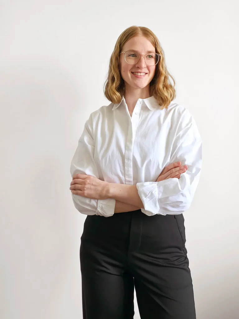 Anika Schulze, Freelance Grafikdesignerin aus Hamburg. Sie hat schulterlanges, lockiges, blondes Haar und trägt eine rosa, durchsichtige Brille. Sie steht vor einer wie-en Wand und trägt eine weisse Hemdbluse und eine schwarze Anzughose. Ihre Arme sind auf Hüfthöhe verschränkt und sieht lächelt. Mit Studio Schulze bietet sie Branding & Webdesign für selbstständige Frauen an.