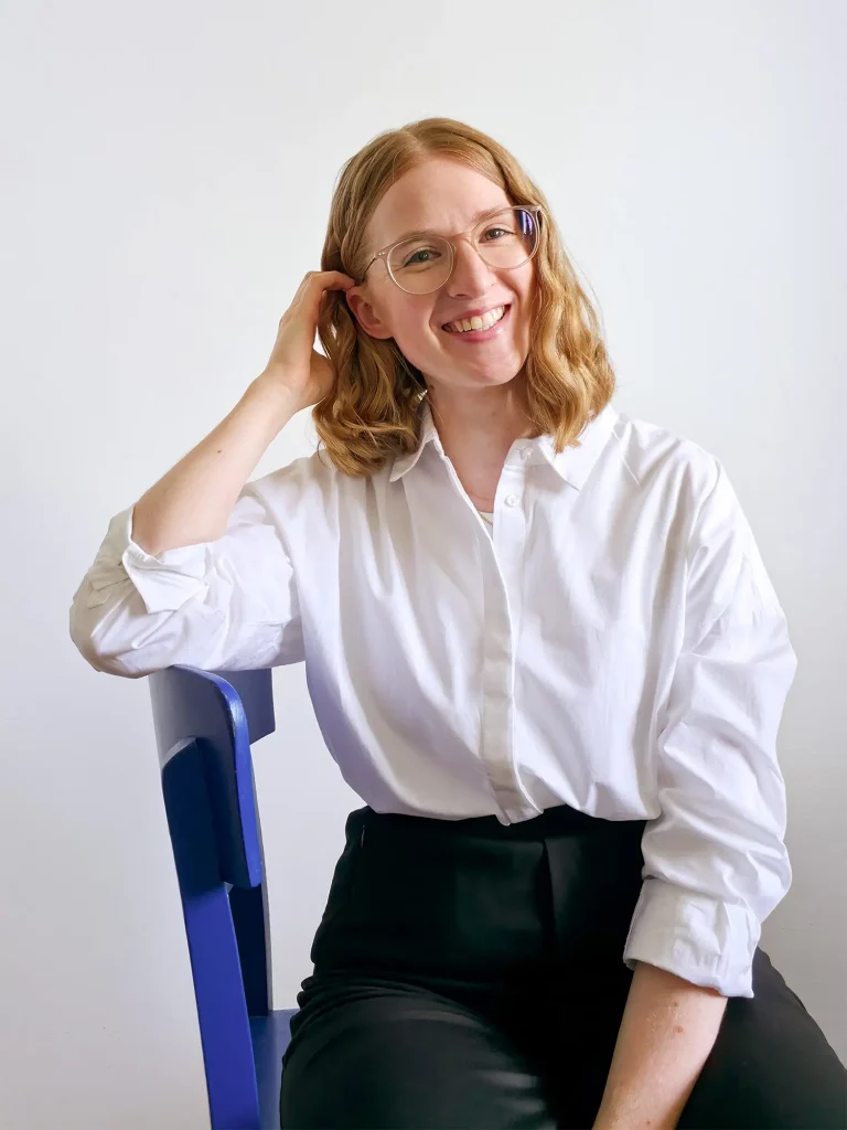 Anika Schulze, Freelance Brand Designerin aus Hamburg. Sie hat schulterlanges, lockiges, blondes Haar und trägt eine rosa, durchsichtige Brille. Sie steht vor einer wie-en Wand und trägt eine wie-e Hemdbluse und eine schwarze Anzughose.