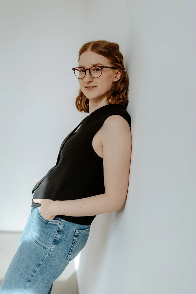 Grafikdesignerin mit Brille lehnt seitlich an einer weißen Wand, Hände in den Hosentaschen, schwarzes Oberteil und Jeans.