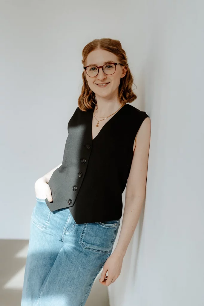 Grafikdesignerin mit Brille lehnt an einer weißen Wand, trägt schwarze Weste und Jeans und lächelt in die Kamera.