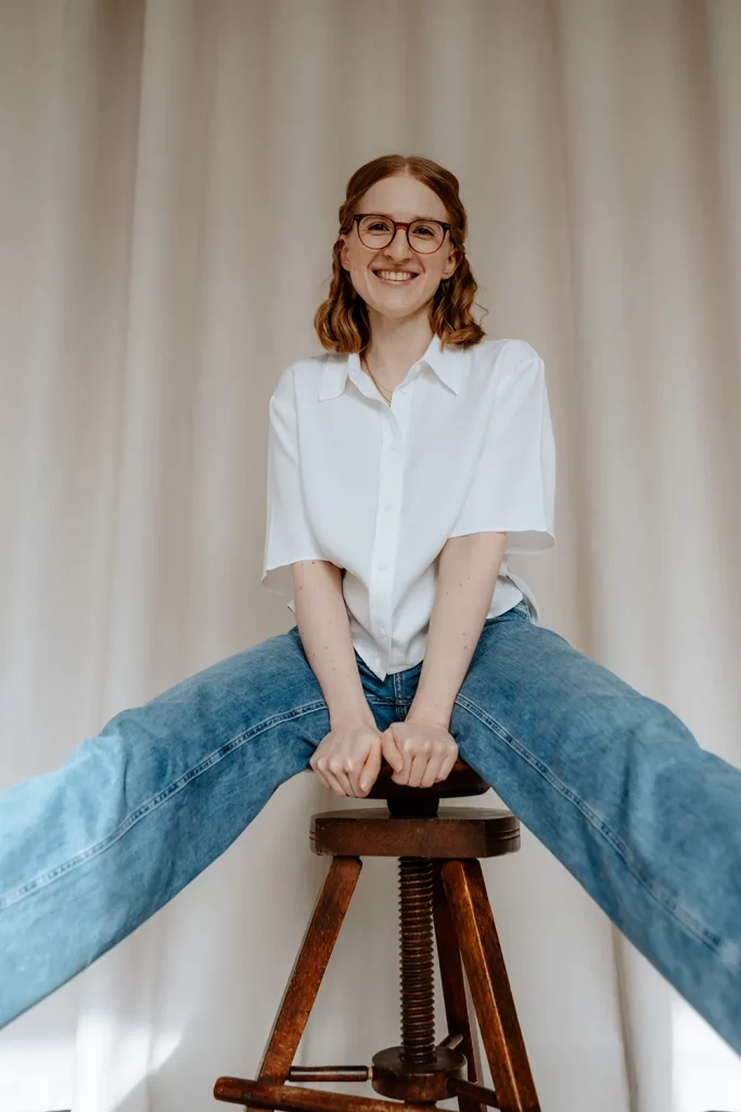 Person, Anika Schulze Grafikdesignerin, mit Brille sitzt breitbeinig auf einem Hocker und lächelt in die Kamera, weißes Hemd und Jeans vor hellem Vorhang.
