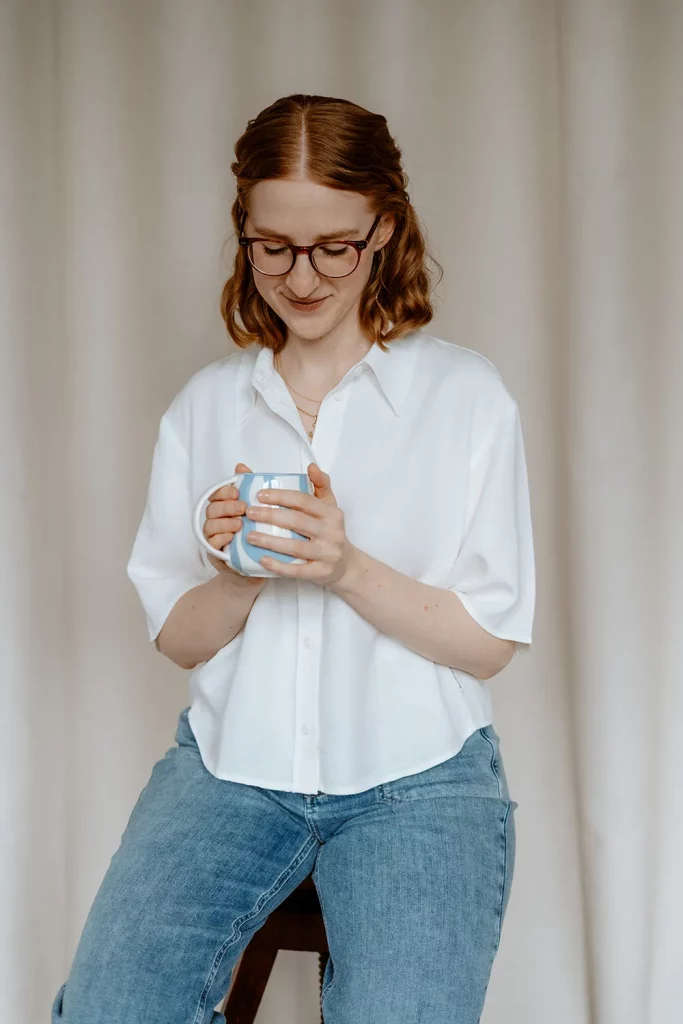 Person, Anika Schulze Grafikdesignerin, mit Brille sitzt auf einem Hocker, schaut auf eine Tasse in den Händen, weißes Hemd und Jeans vor hellem Vorhang.
